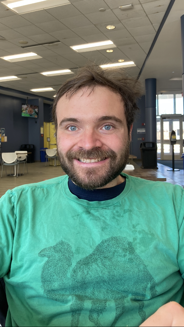 Photograph shows a man with brown hair and a beard wearing a bright green shirt featuring camel. He is indoors with tables and chairs in the background.