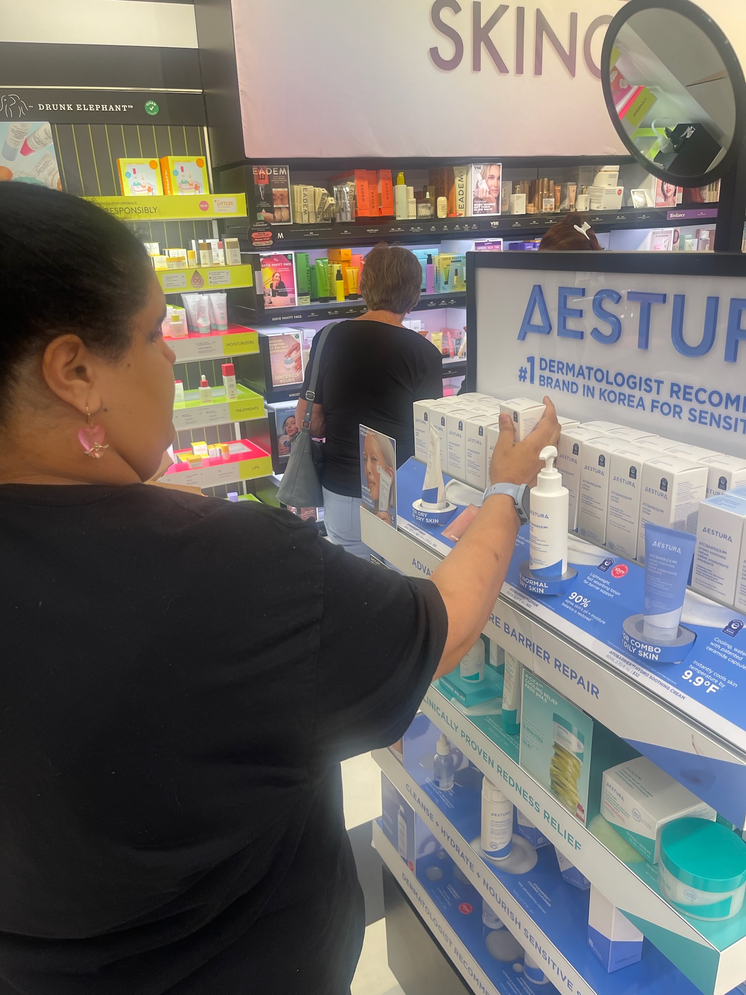 Photograph of a skincare store display, showing a Emely examining Aestura products beside shelves stocked with colorful cosmetics and personal-care items.