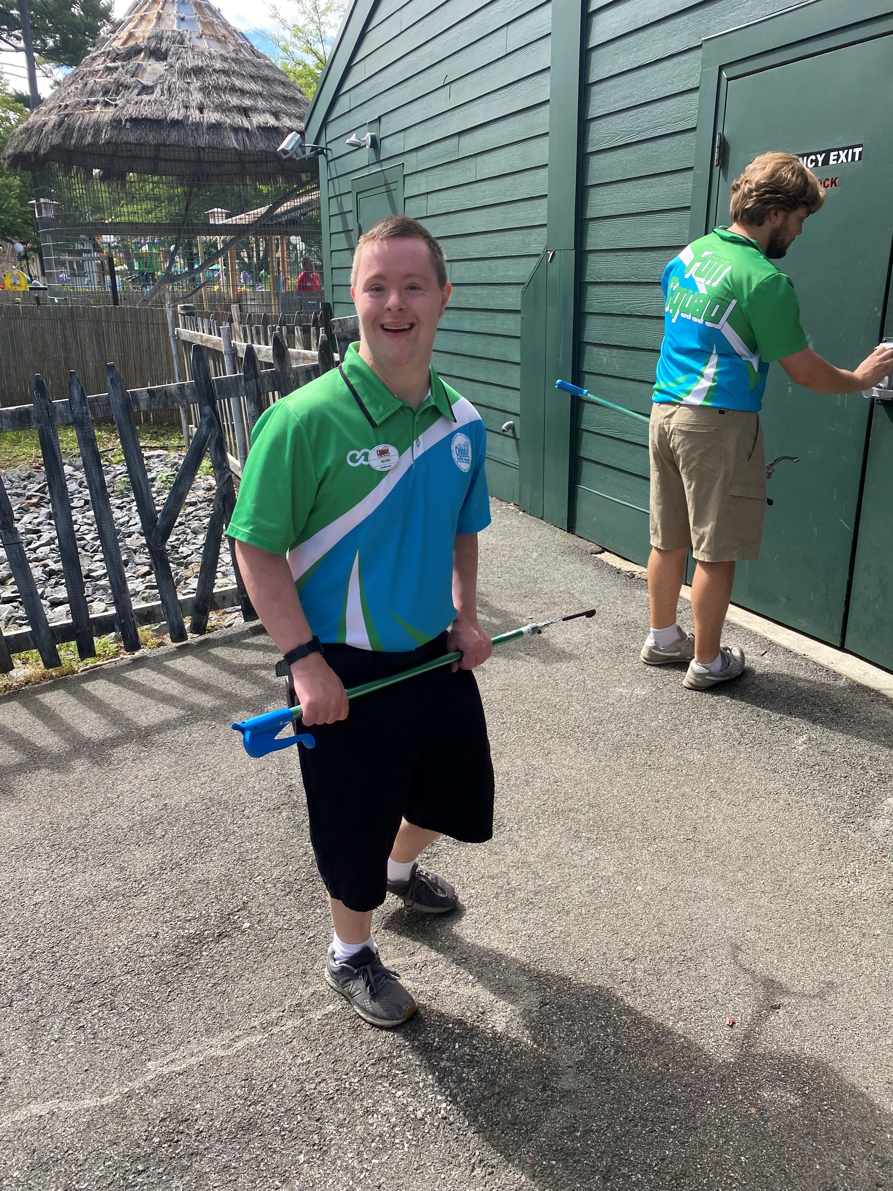 Photograph shows smiling man in green-and-blue polo holding a long metal item. Another person opens an EXIT door.