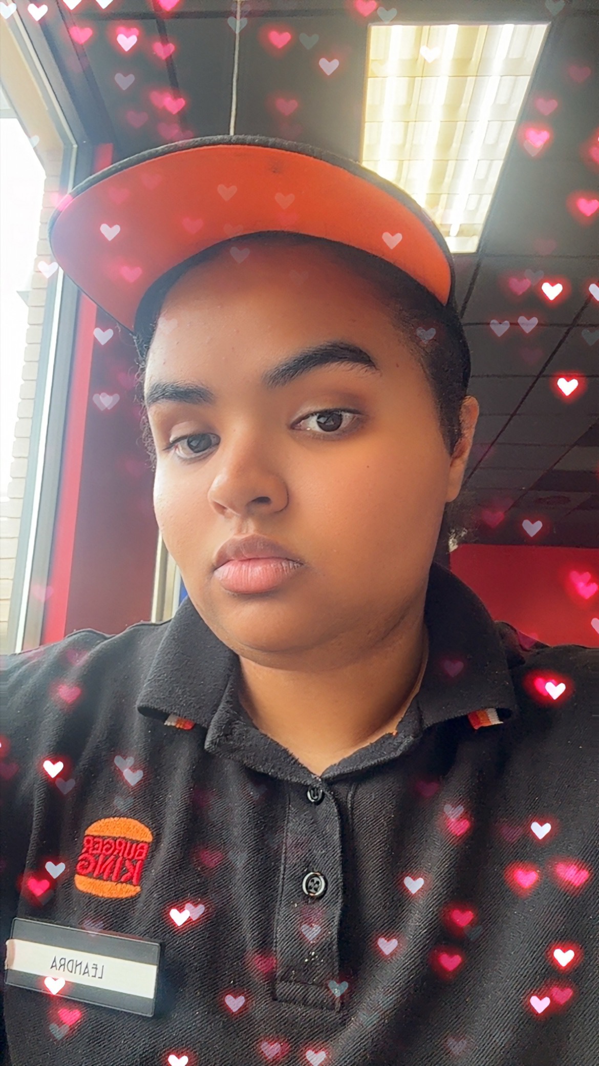 Photograph shows Leandra taking a selfie indoors while wearing a red cap and black collared work uniform with a name badge. Pink and red heart graphics overlay face and restaurant-like background.